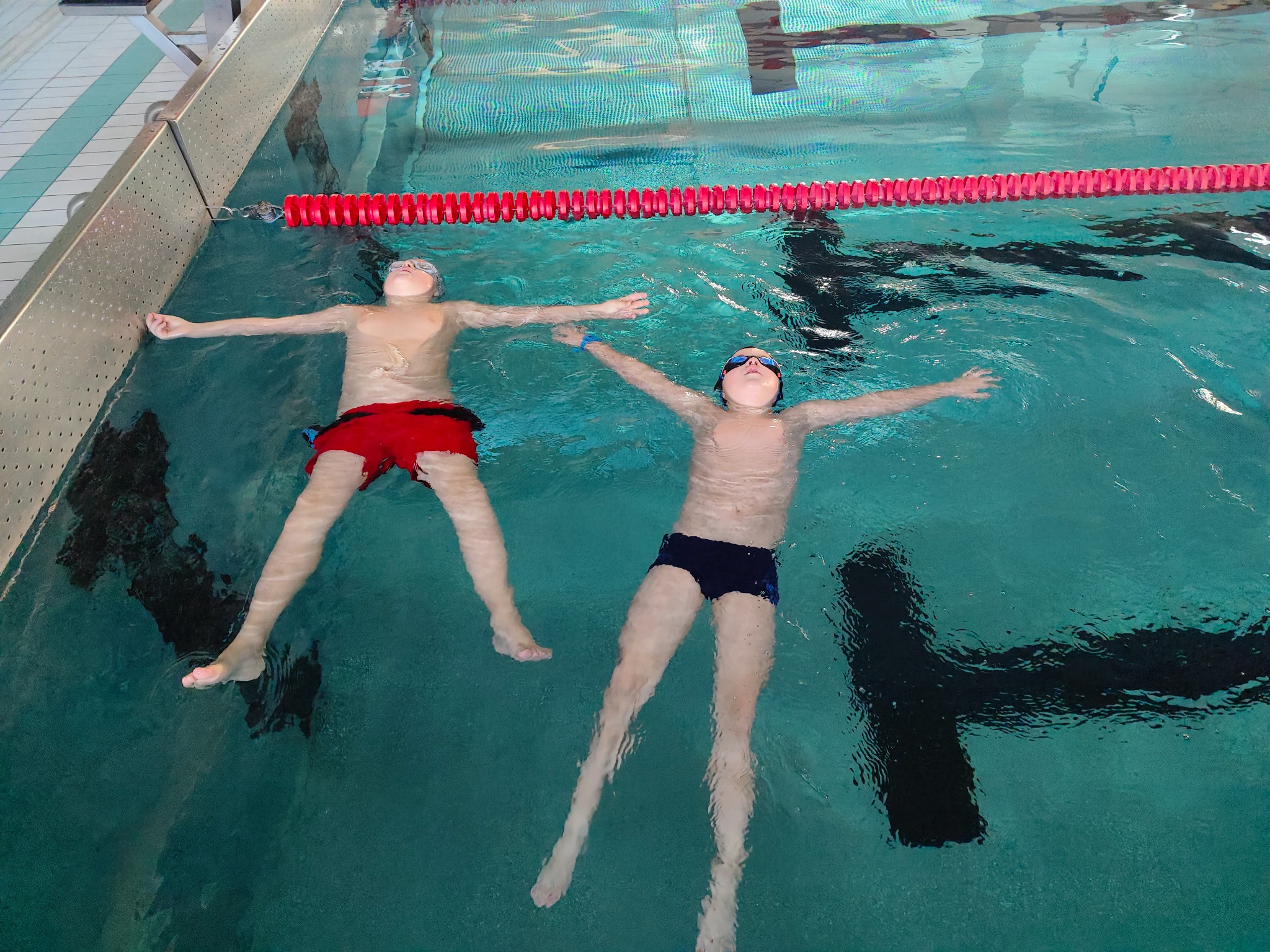 Dwaj chłopcy w okularach pływackich unoszą się na wodzie w basenie, z rozłożonymi ramionami, blisko czerwonej liny oddzielającej tory.
