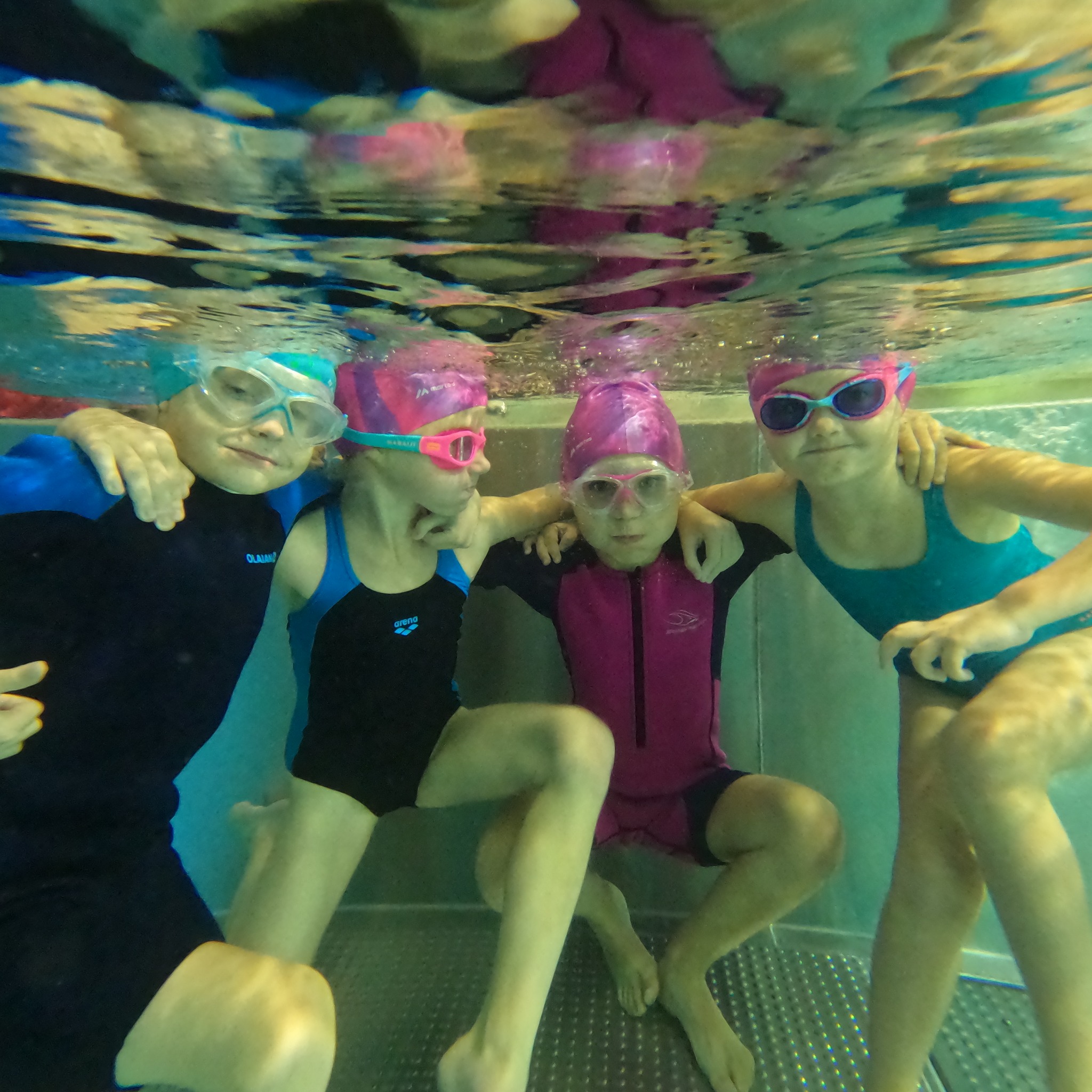 Cztery uśmiechnięte dziewczynki w czepkach i okularach pływackich, zanurzone w basenie, obejmują się pod wodą. Widok od dołu, z perspektywy osoby znajdującej się pod nimi.