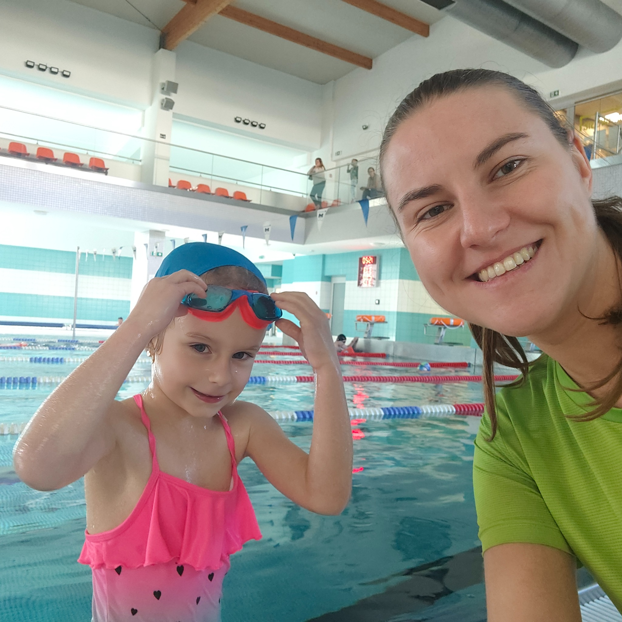 Instruktorka z uśmiechem robi selfie z dziewczynką w czepku i okularkach na basenie. W tle woda i osoby korzystające z pływalni.