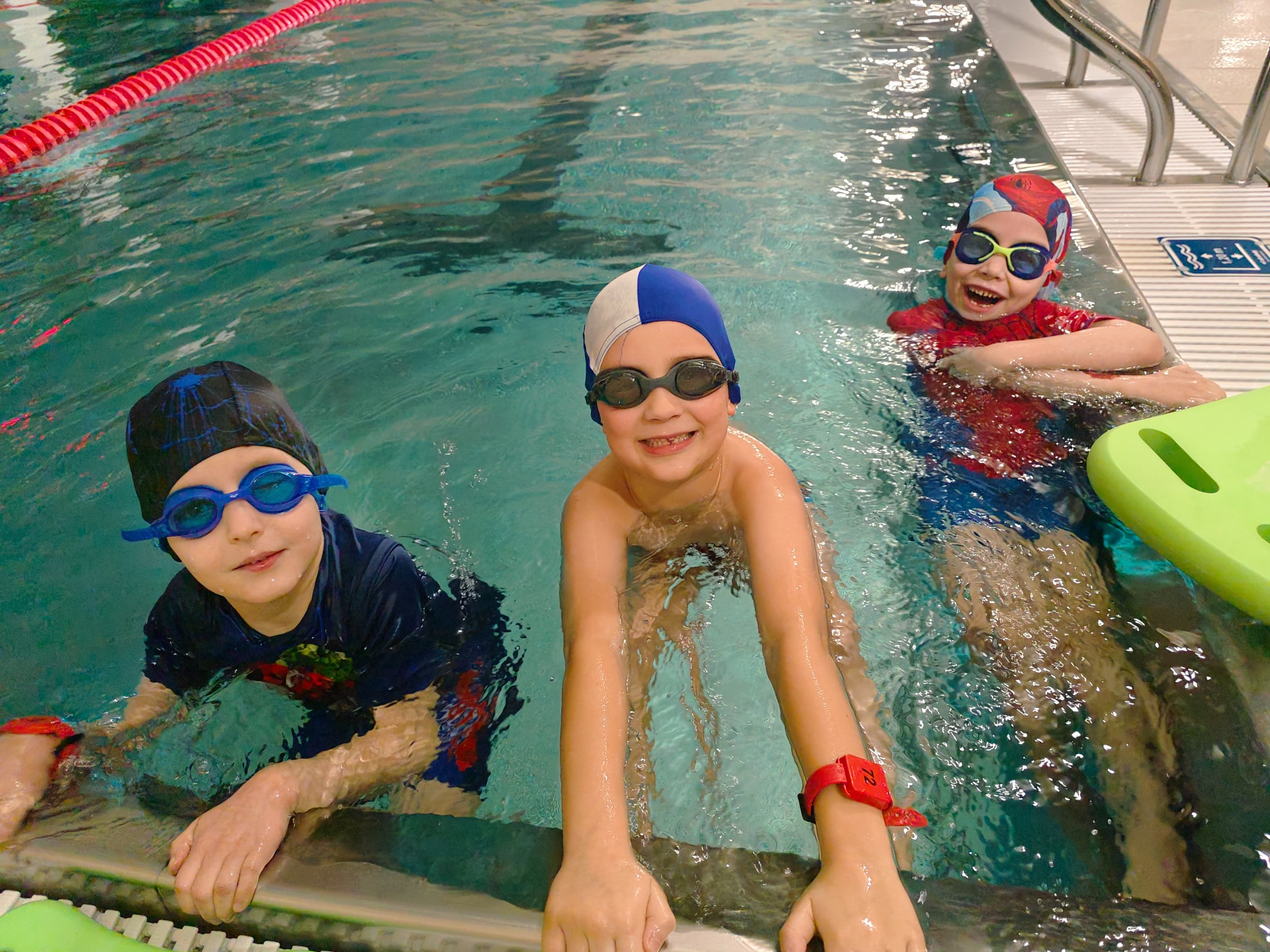 Trójka uśmiechniętych dzieci w czepkach i okularach pływackich w basenie, z czerwonymi opaskami na nadgarstkach, opierających się o brzeg. W tle widoczny tor pływacki.