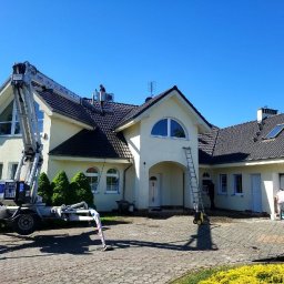 AlpinClean - Dwupiętrowy dom z beżową elewacją podczas mycia. Na dachu i przy elewacji pracownicy z użyciem drabiny i podnośnika koszowego. Kostka brukowa mokra po myciu.