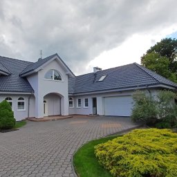 AlpinClean - Jasnoszary dom z ciemnym dachem i kostką brukową na podjeździe, otoczony zielenią. Widok z poziomu gruntu, pochmurne niebo w tle.