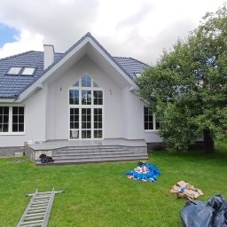 AlpinClean - Biała elewacja domu jednorodzinnego po malowaniu, z widoczną drabiną i pracownikiem. Na trawniku akcesoria malarskie, tło: niebo z chmurami i drzewo.