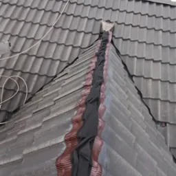 Szczegółowe ujęcie dachu z brązową powłoką i czarnym zabezpieczeniem na łączeniu połaci, antena na szczycie. Widok z góry, pochmurne niebo w tle.