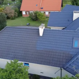 Mycie dachu z szarej dach&oacute;wki z wyraźną r&oacute;żnicą czystości, widok z g&oacute;ry na domy i zieleń w tle, fragment metalowej konstrukcji w rogu kadru.