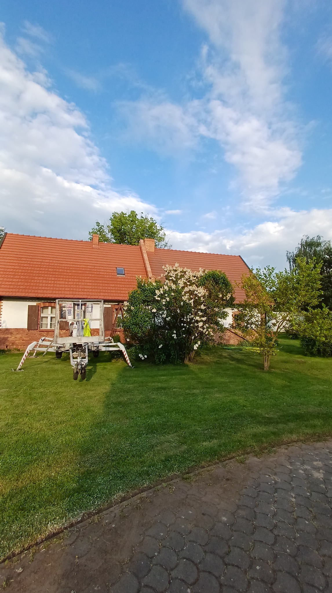 Podnośnik koszowy na trawniku przed domem z czerwoną dachówką i kwitnącym krzewem. Błękitne niebo z chmurami w tle.