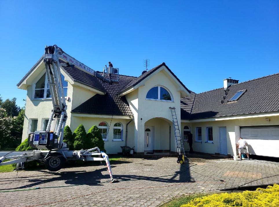 Dwupiętrowy dom z beżową elewacją podczas mycia. Na dachu i przy elewacji pracownicy z użyciem drabiny i podnośnika koszowego. Kostka brukowa mokra po myciu.