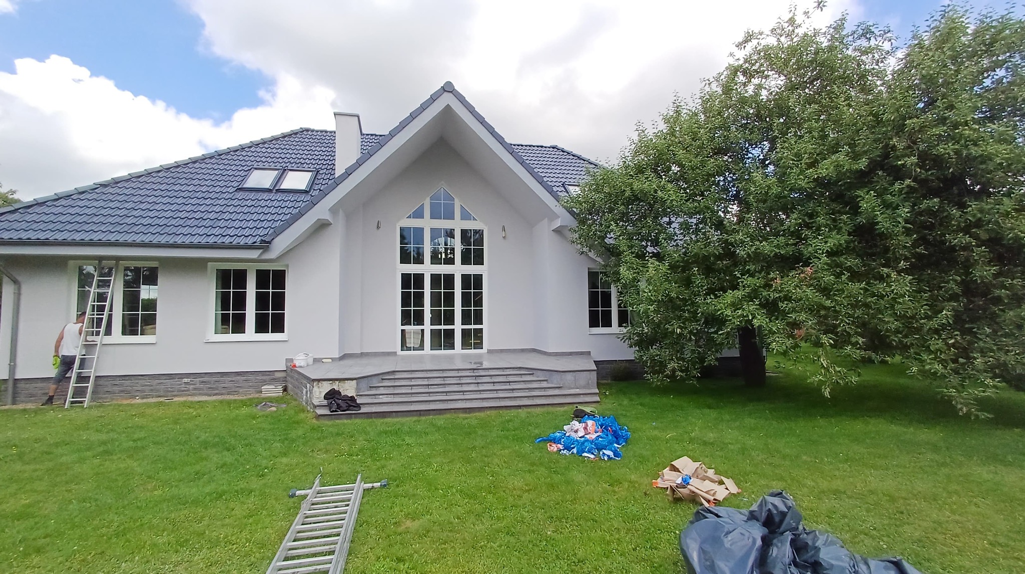Biała elewacja domu jednorodzinnego po malowaniu, z widoczną drabiną i pracownikiem. Na trawniku akcesoria malarskie, tło: niebo z chmurami i drzewo.