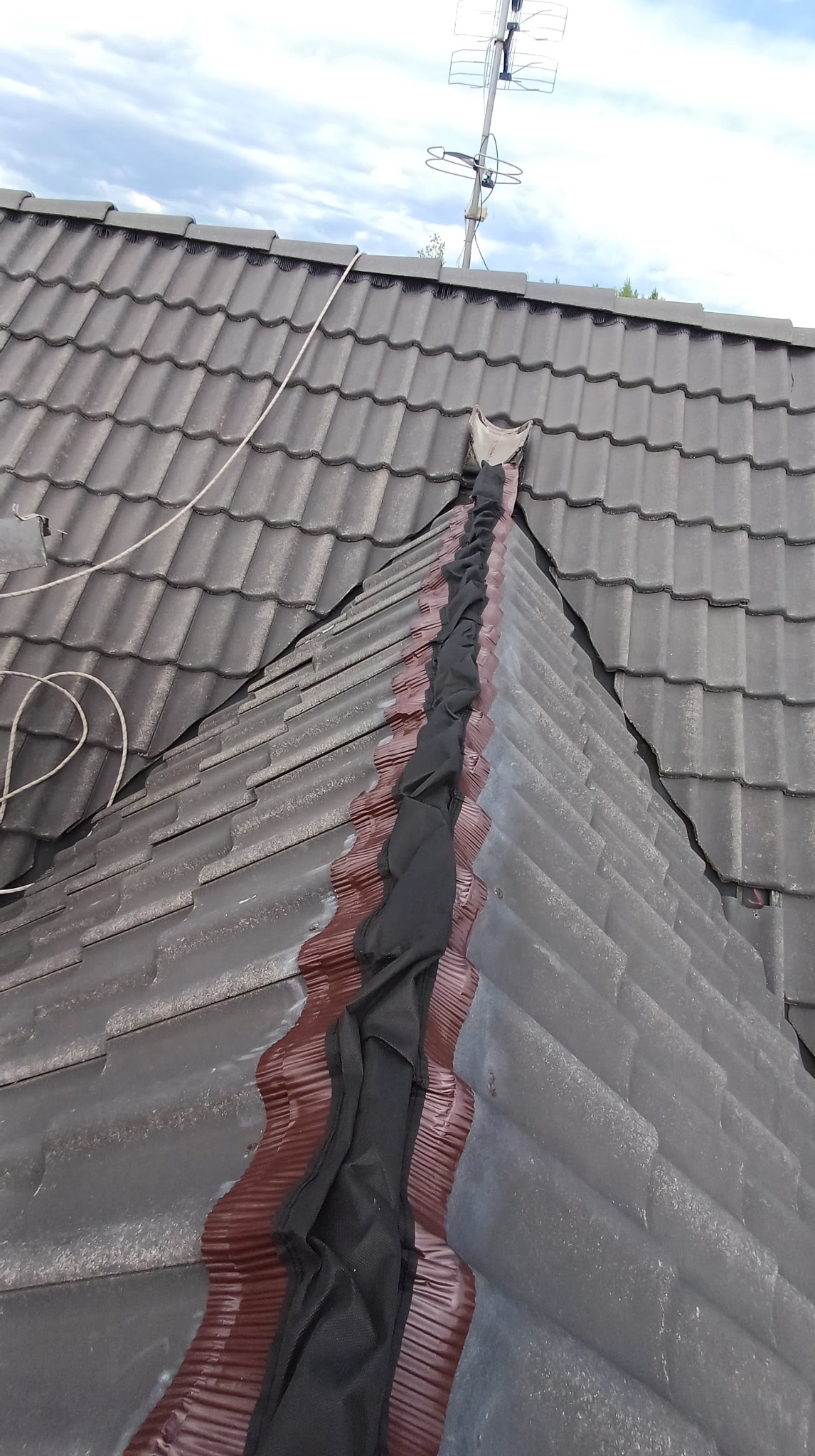 Szczegółowe ujęcie dachu z brązową powłoką i czarnym zabezpieczeniem na łączeniu połaci, antena na szczycie. Widok z góry, pochmurne niebo w tle.