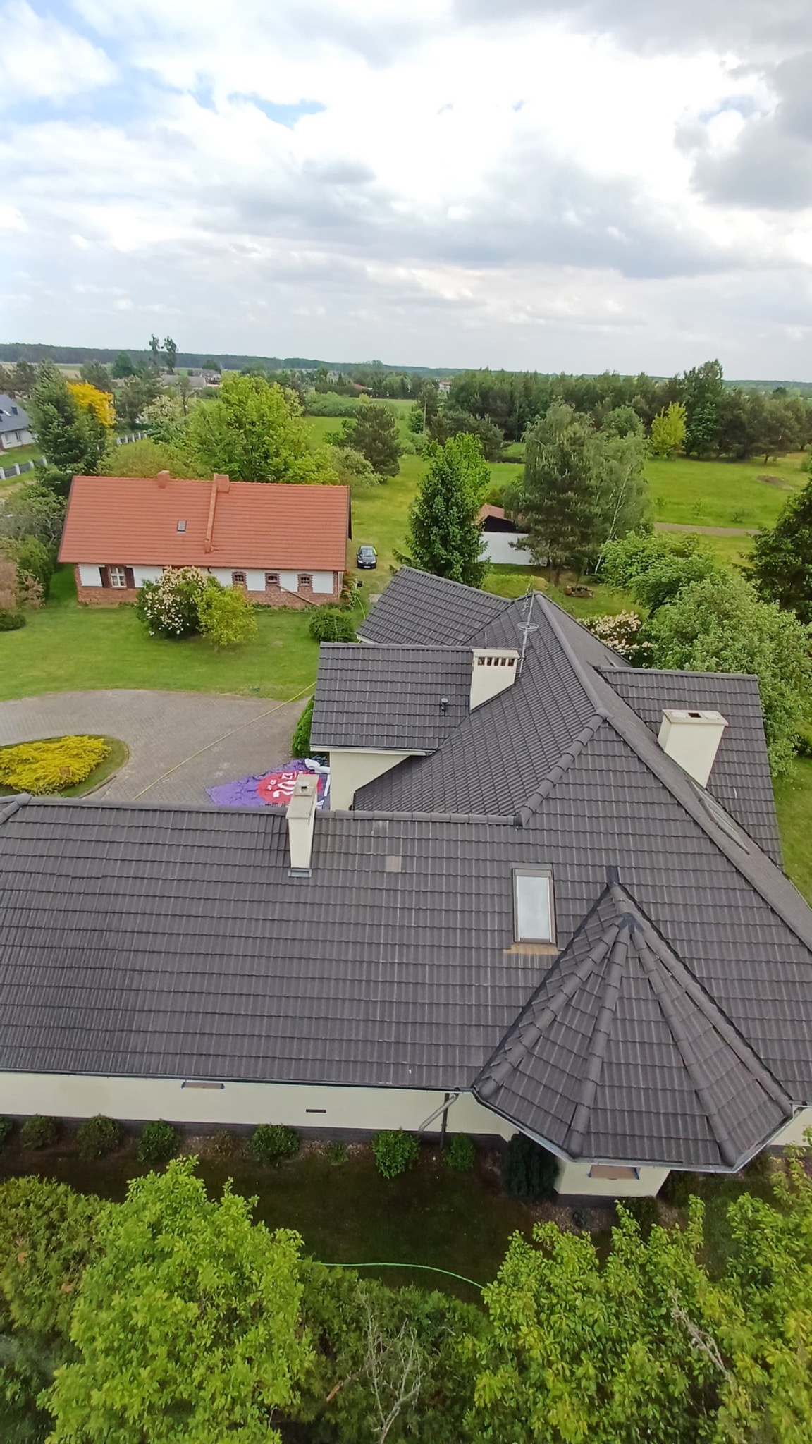 Widok z góry na duży dom z ciemnoszarym dachem, kominami i oknem dachowym. W tle zieleń drzew i trawników, a także drugi dom z czerwonym dachem.