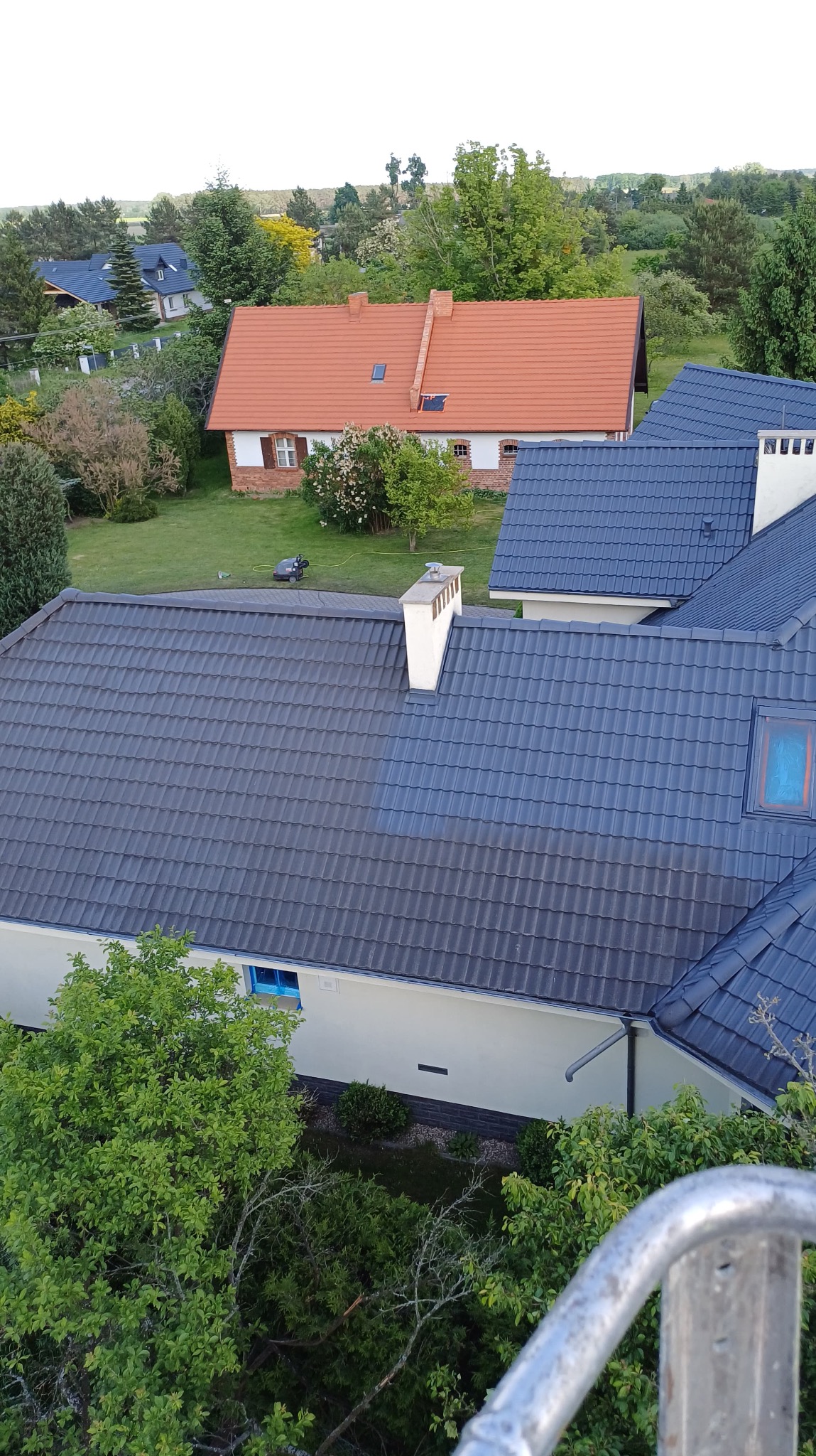 Mycie dachu z szarej dachówki z wyraźną różnicą czystości, widok z góry na domy i zieleń w tle, fragment metalowej konstrukcji w rogu kadru.