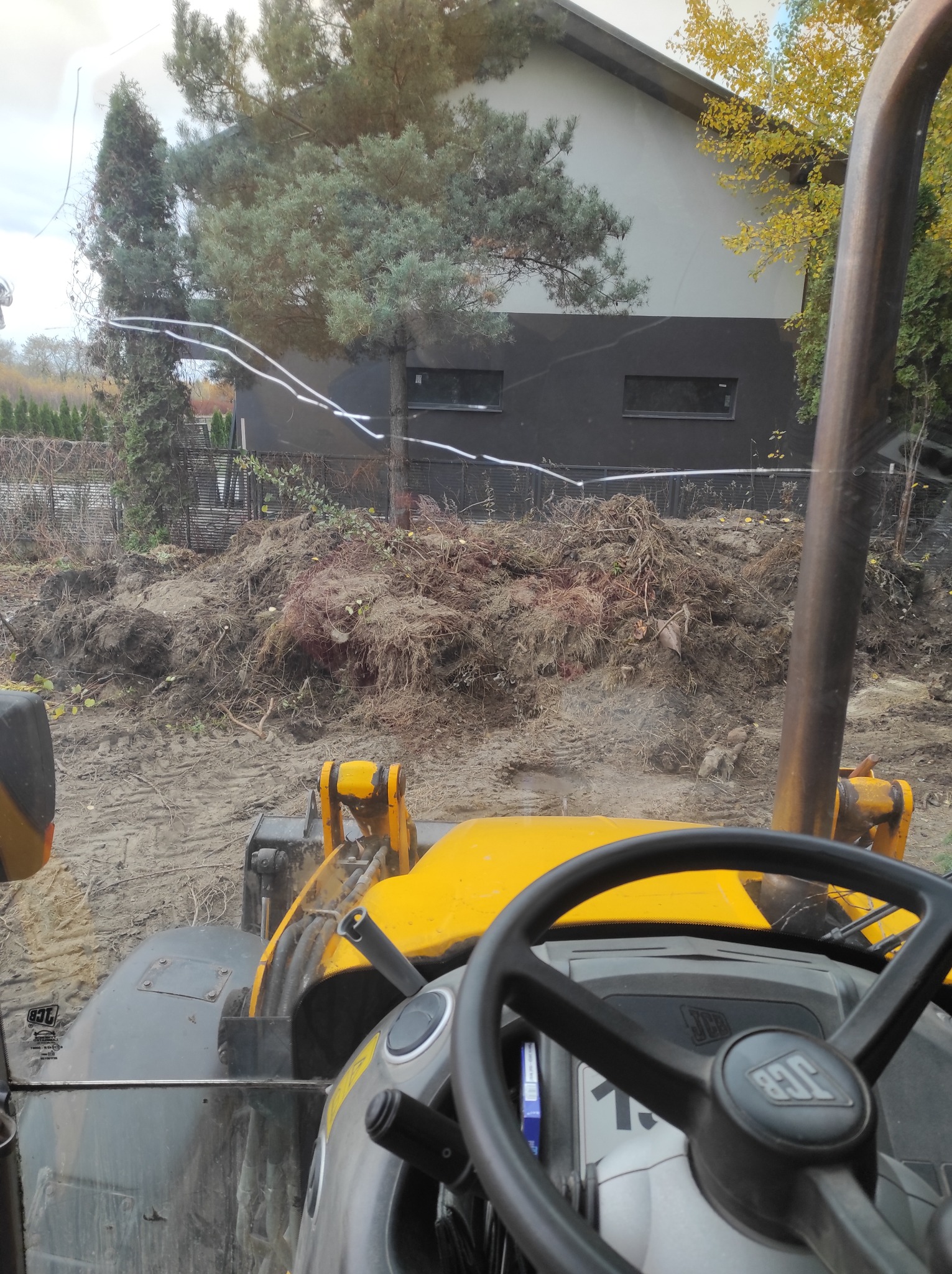 Widok z kabiny żółtej koparki JCB na stertę ziemi i gruzu przed nowoczesnym domem. Ślady opon na ziemi. Praca w toku.