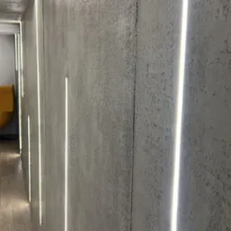 Szara ściana z fakturą betonu i pionowymi, wpuszczanymi paskami LED, oświetlającymi korytarz. Widoczne drewnopodobne płytki na podłodze i fragment żółtego fotela.