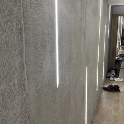 Thermobud Sp. z o.o. - Szara ściana z fakturą i pionowymi pasami światła LED, oświetlającymi korytarz z drewnianą podłogą i butami w tle. Nowoczesny design wnętrza.