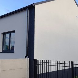 Thermobud Sp. z o.o. - Elewacja domu po malowaniu w Mikołowie, z jasnymi ścianami i ciemnymi detalami, widok z poziomu ogrodzenia, w tle budynek z cegły.