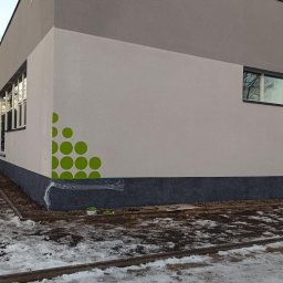 Thermobud Sp. z o.o. - Wykończona elewacja budynku z dekoracyjnym, zielonym wzorem w postaci kół. Widoczne okna i fragment ośnieżonego terenu. Detal wykończenia.