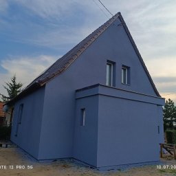 Thermobud Sp. z o.o. - Dom jednorodzinny z nową, gładką elewacją w odcieniach szarości, widoczny z perspektywy bocznej. Dach pokryty brązową dachówką. Widoczne puszki z farbą.