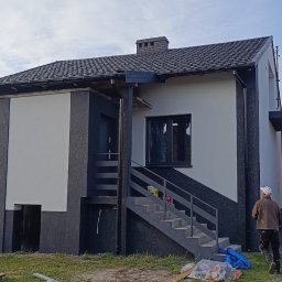 Thermobud Sp. z o.o. - Dwukondygnacyjny dom z nową elewacją w odcieniach szarości i bieli, z widocznymi schodami zewnętrznymi i pracownikiem w tle. Częściowo widoczne narzędzia i farby.