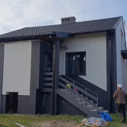 Dwukondygnacyjny dom z nową elewacją w odcieniach szarości i bieli, z widocznymi schodami zewnętrznymi i pracownikiem w tle. Częściowo widoczne narzędzia i farby.