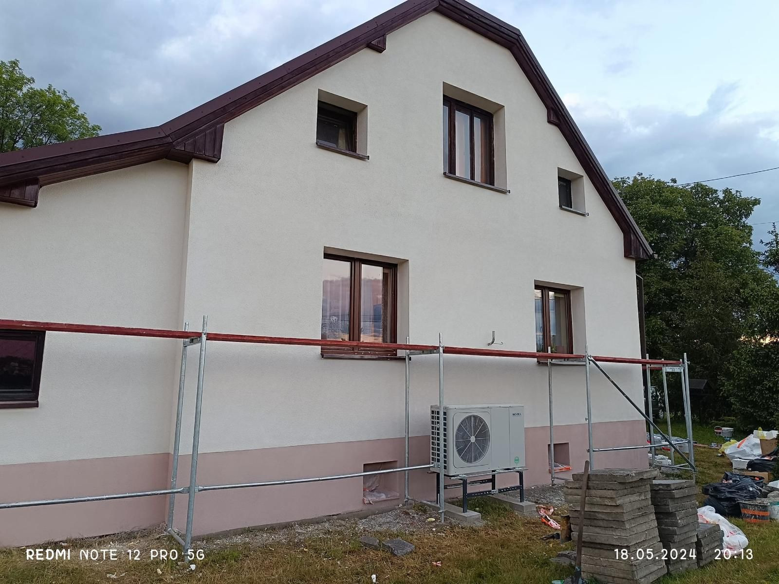 Jasna elewacja domu z brązowym dachem, widoczne rusztowanie i klimatyzator. Dolna część elewacji pomalowana na różowo. Widok z boku, wieczorne światło.