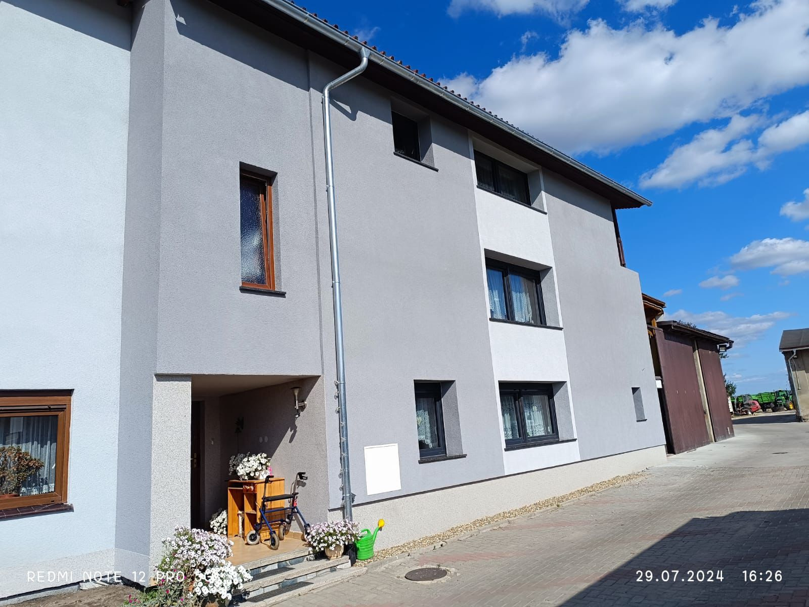 Odnowiona elewacja domu w odcieniach szarości, z jasnym akcentem przy oknach. Widoczne kwiaty i elementy dekoracyjne przy wejściu. Błękitne niebo w tle.