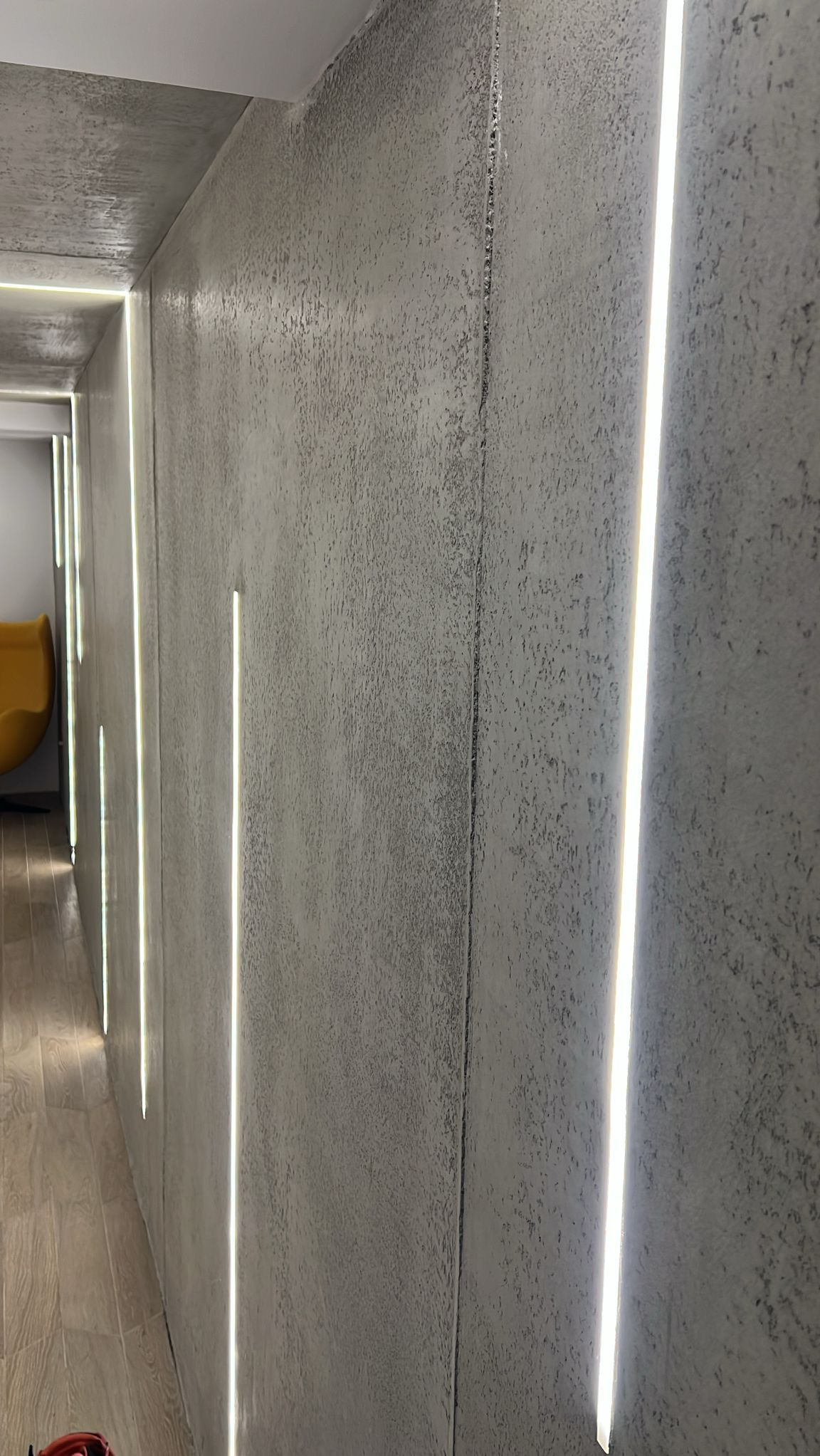 Szara ściana z fakturą betonu i pionowymi, wpuszczanymi paskami LED, oświetlającymi korytarz. Widoczne drewnopodobne płytki na podłodze i fragment żółtego fotela.