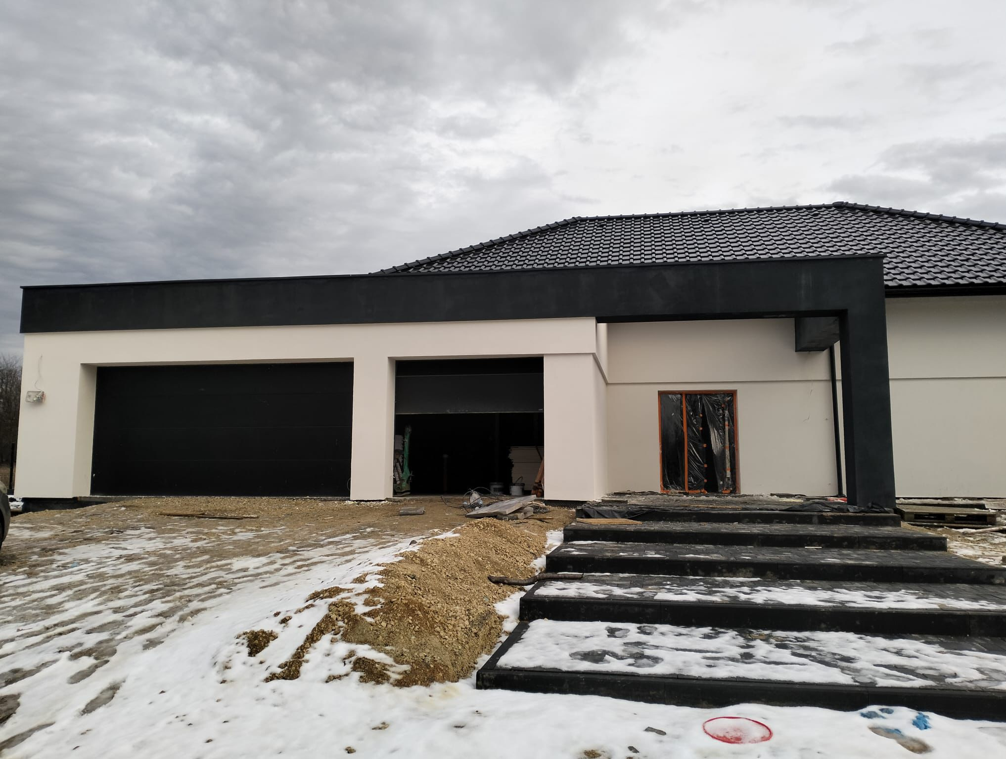Nowoczesny dom z garażem i czarnymi schodami wejściowymi. Elewacja w odcieniach bieli i szarości, czarny dach. Wokół resztki śniegu. Widok na front budynku.