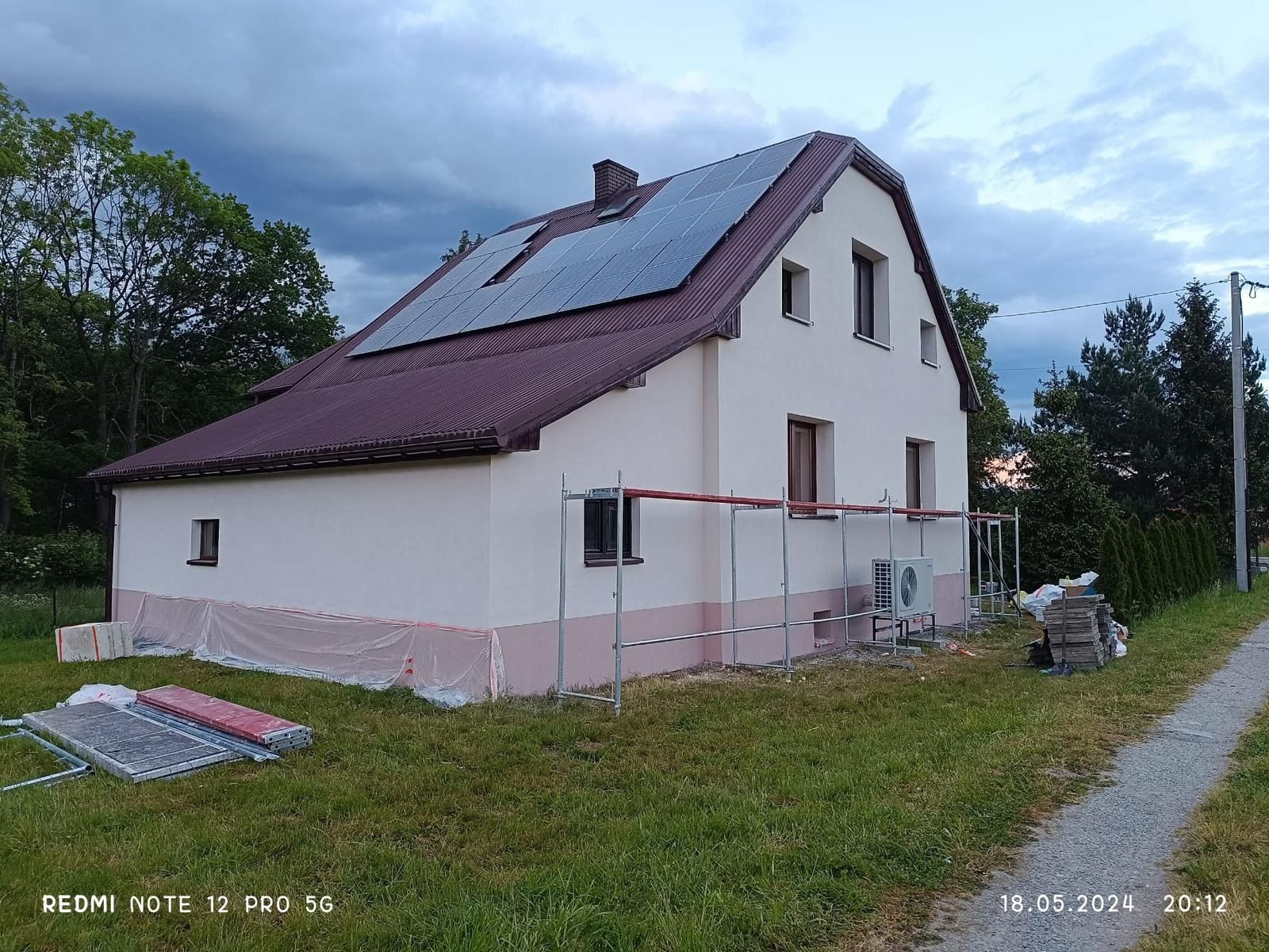 Dom z panelami słonecznymi na dachu, częściowo osłonięty rusztowaniem. Widoczna klimatyzacja przy ścianie. Elewacja dwukolorowa, jasna góra i różowy dół.