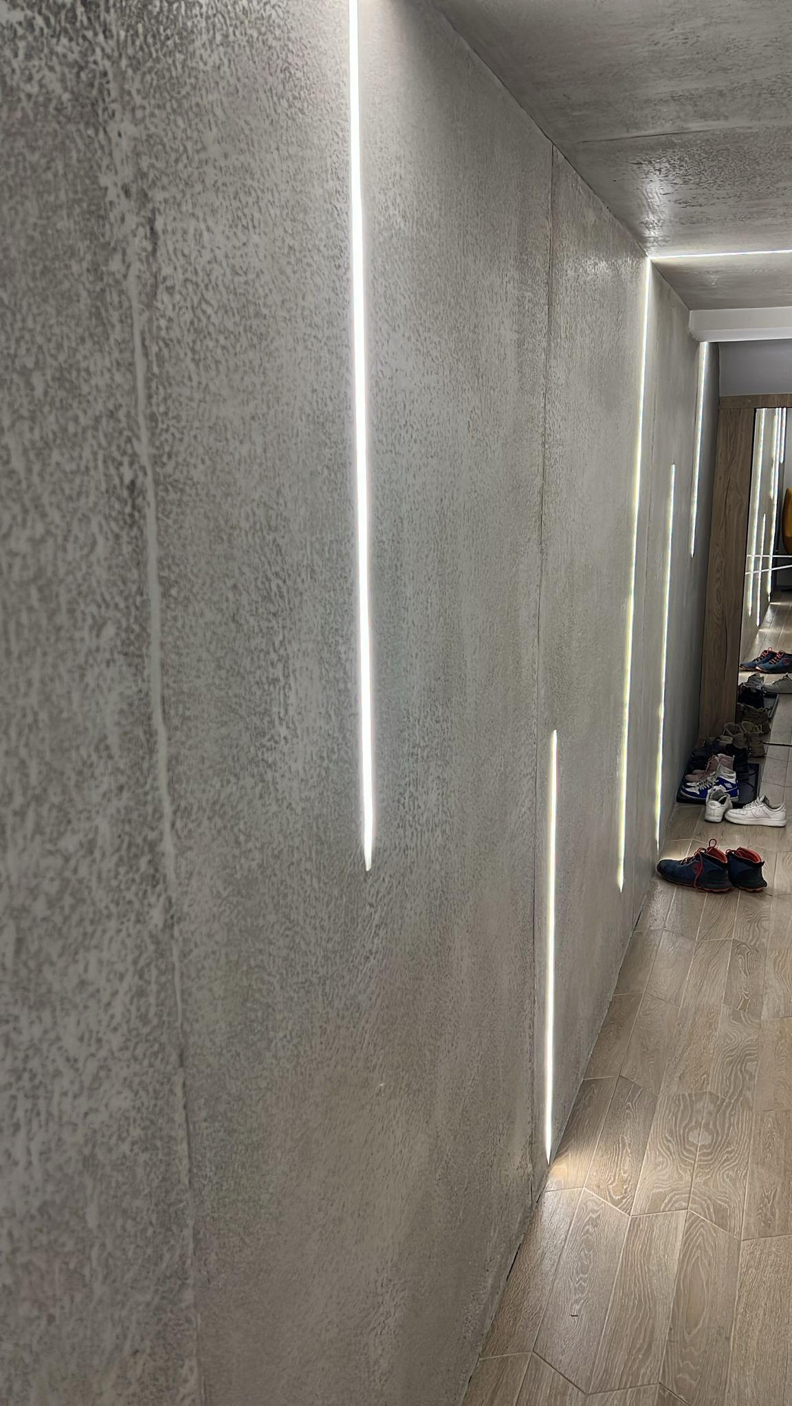 Szara ściana z fakturą i pionowymi pasami światła LED, oświetlającymi korytarz z drewnianą podłogą i butami w tle. Nowoczesny design wnętrza.