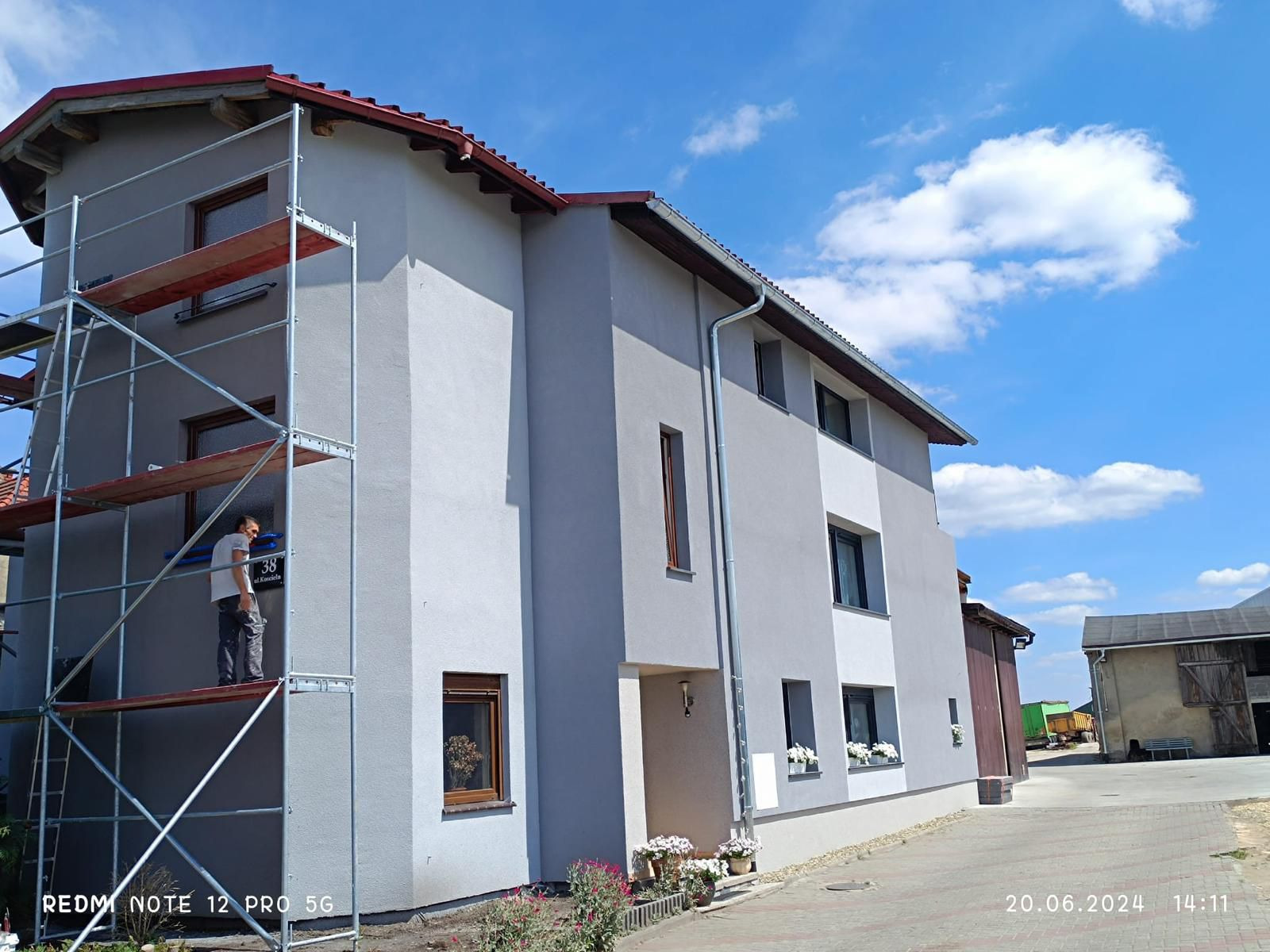 Dwukondygnacyjny dom z szarą elewacją, częściowo pokrytą rusztowaniem, na którym pracuje mężczyzna. Widoczne kwiaty i fragment chodnika.