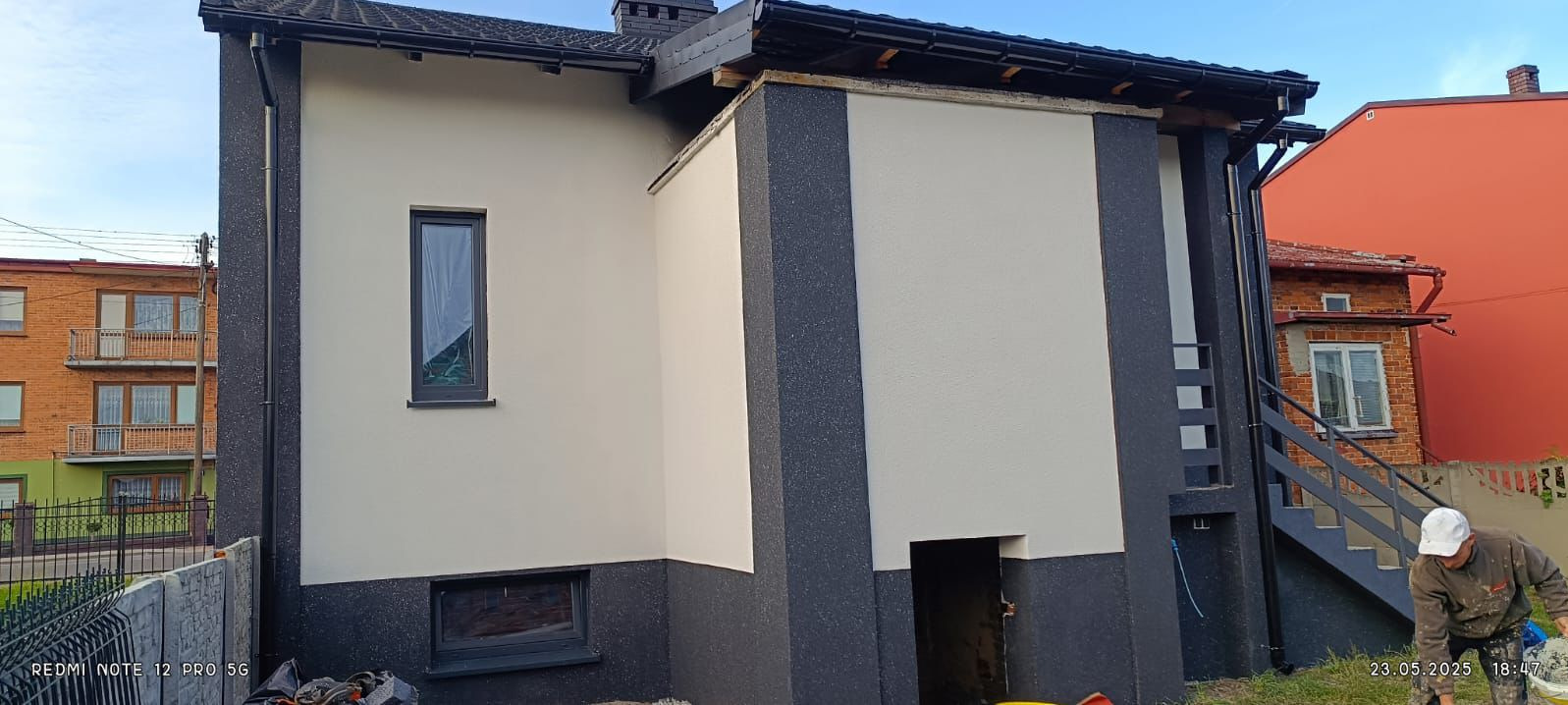 Dwukolorowa elewacja domu jednorodzinnego, z elementami w odcieniach szarości i jasnego beżu, widok z zewnątrz, częściowo widoczny pracownik w trakcie pracy.