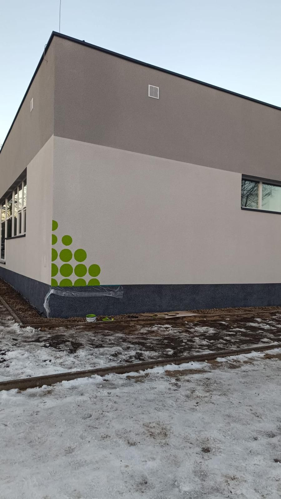 Wykończona elewacja budynku z dekoracyjnym, zielonym wzorem w postaci kół. Widoczne okna i fragment ośnieżonego terenu. Detal wykończenia.