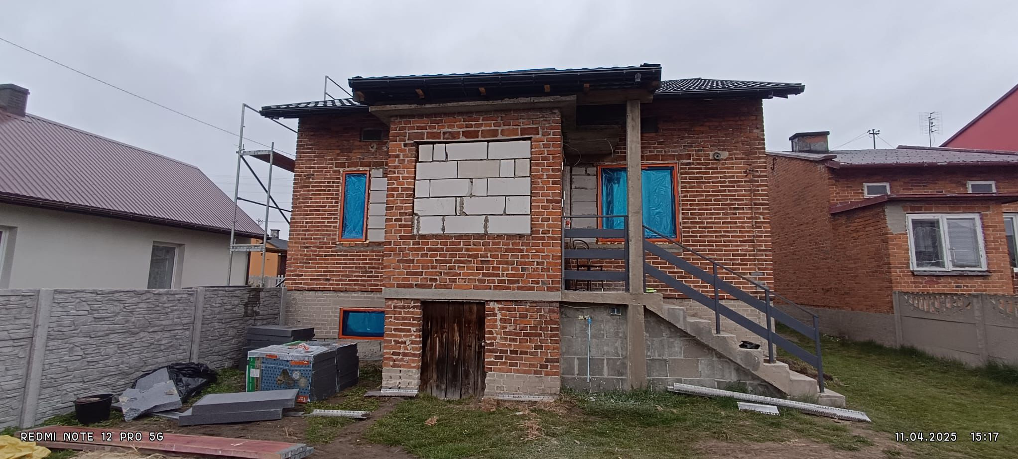 Ceglany dom w trakcie prac elewacyjnych: okna zabezpieczone folią, częściowo otynkowana ściana, widoczne materiały budowlane, rusztowanie z boku.