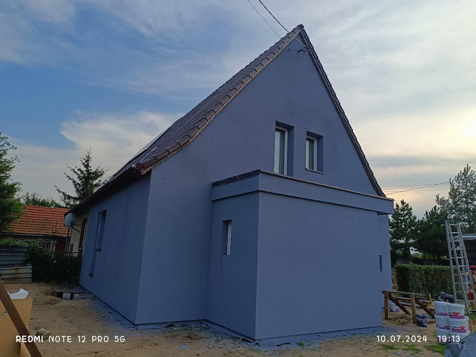 Dom jednorodzinny z nową, gładką elewacją w odcieniach szarości, widoczny z perspektywy bocznej. Dach pokryty brązową dachówką. Widoczne puszki z farbą.