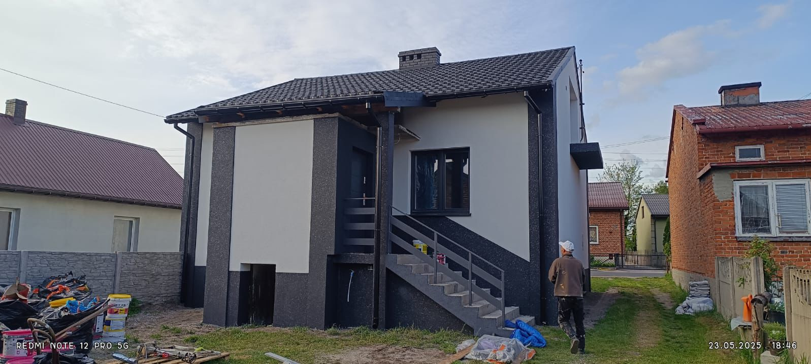 Dwukondygnacyjny dom z nową elewacją w odcieniach szarości i bieli, z widocznymi schodami zewnętrznymi i pracownikiem w tle. Częściowo widoczne narzędzia i farby.