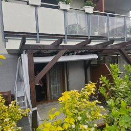 N8 Struct - Drewniana pergola z roletą i zasłonami na tarasie, widok z ogrodu. Na balkonie nad nią donice z kwiatami, a obok drabina oparta o ścianę.