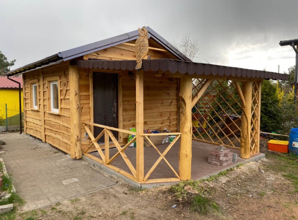 Drewniany domek letniskowy z rzeźbą sowy na dachu werandy, wykończony w naturalnym stylu, z widoczną konstrukcją z bali i ażurową balustradą.