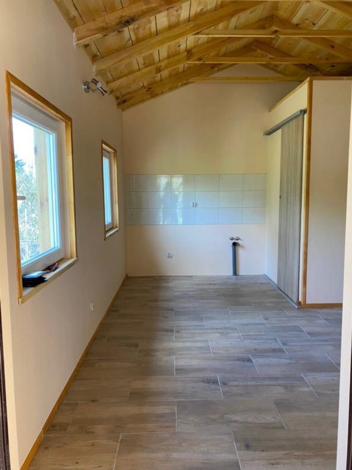 Jasne wnętrze z drewnianą podłogą i skośnym sufitem wykończonym boazerią, okna z drewnianymi ramami, białe płytki na ścianie, widoczna instalacja wodna.