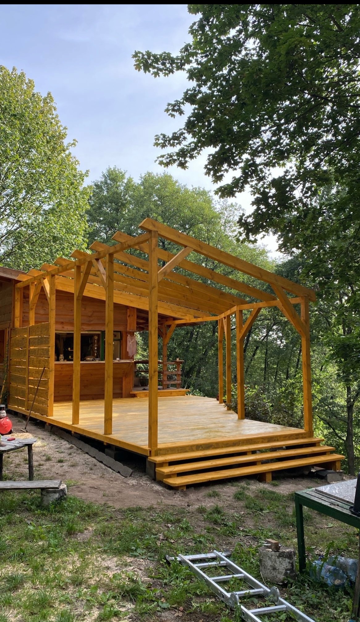 Drewniana konstrukcja altany z podestem i schodami, świeżo pomalowana na żółto, na tle zieleni drzew. Widoczne narzędzia i drabina sugerują trwające prace wykończeniowe.