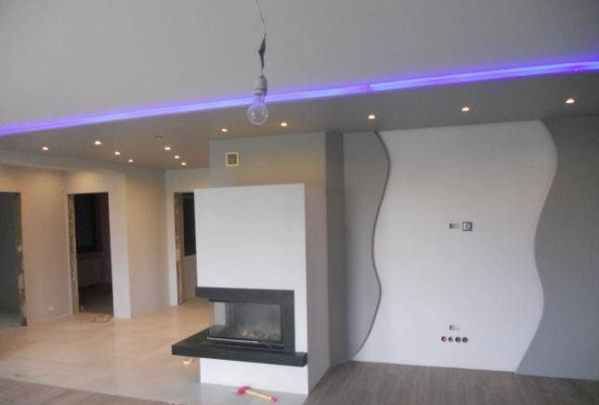 Nowoczesny salon z kominkiem i dekoracyjną ścianą w odcieniach szarości. Sufit podwieszany z oświetleniem LED. Widoczne gniazdka i przełączniki na ścianie.