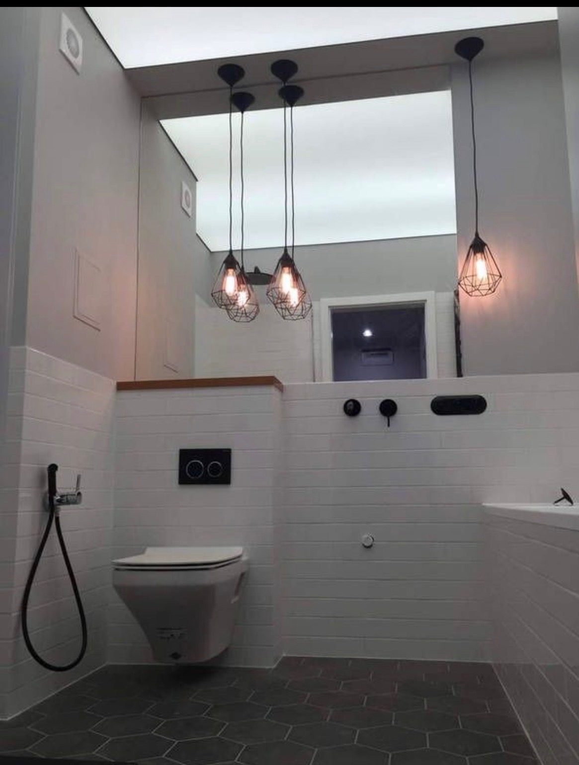 Nowoczesna łazienka z białymi płytkami imitującymi drewno, heksagonalną podłogą i wiszącą toaletą. Cztery lampy z żarówkami Edisona dodają ciepła minimalistycznemu wnętrzu.