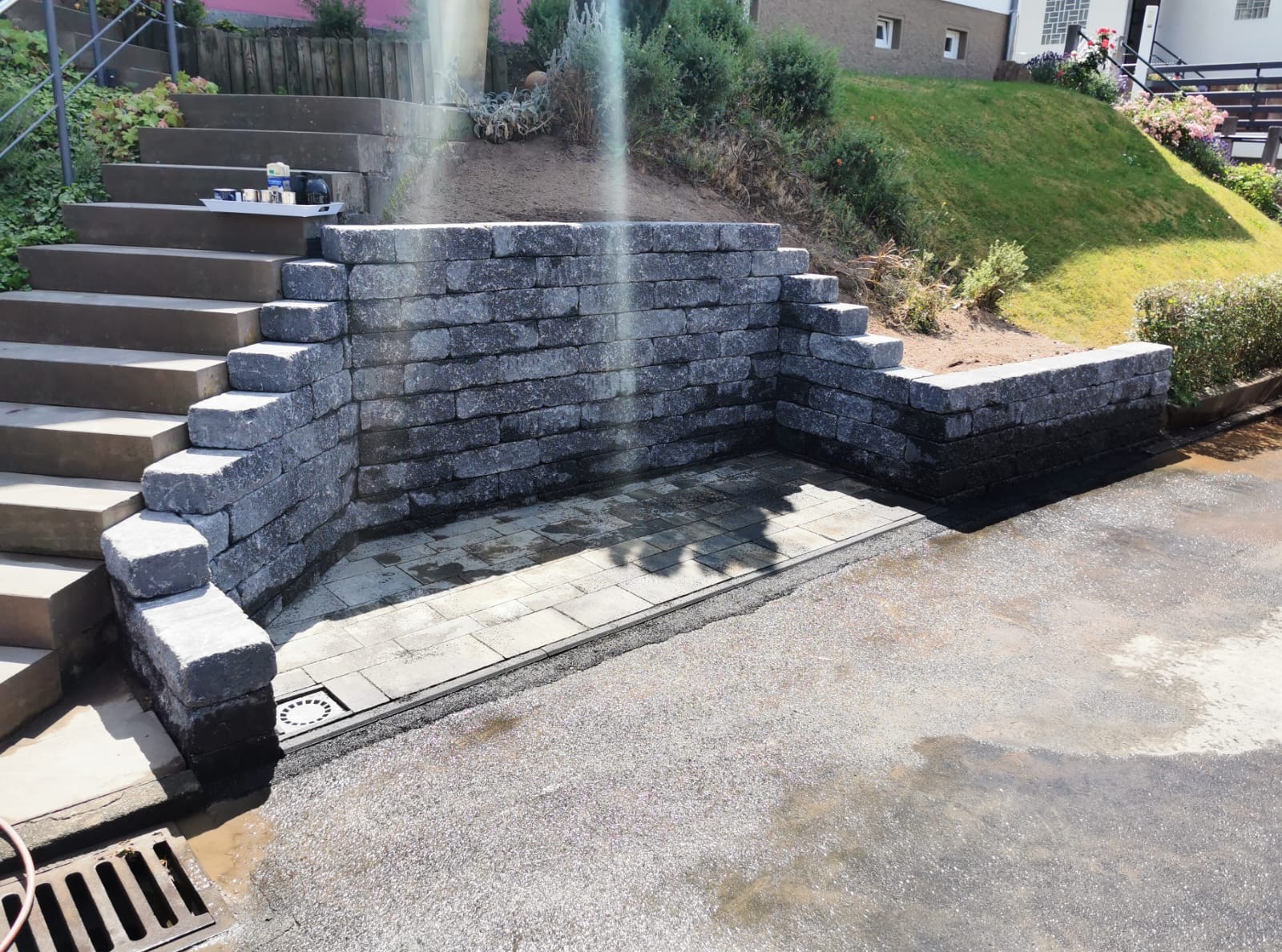 Kamienna ścianka oporowa z kostki brukowej, tworząca mały placyk przy schodach. Widoczne elementy drenażu i mokra nawierzchnia, sugerująca świeżo zakończone prace.