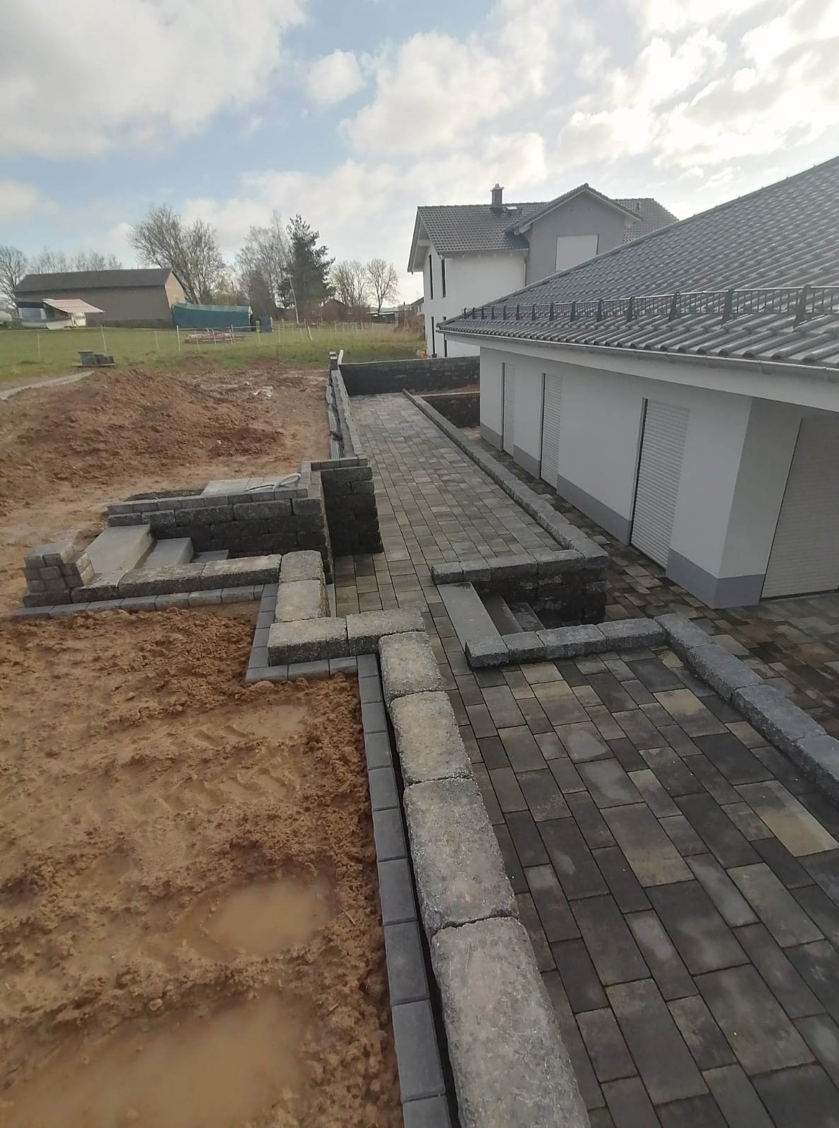 Układanie kostki brukowej na posesji przy domu. Częściowo ułożony chodnik z obrzeżami, ziemia obok. Widok z góry na budynek z szarą dachówką.