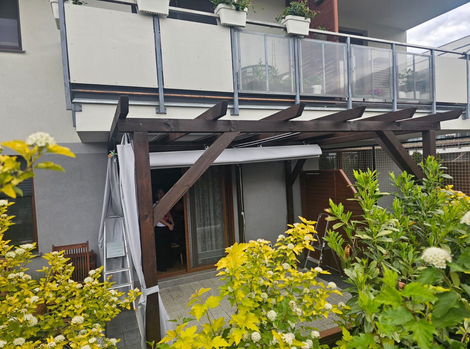 Drewniana pergola z roletą i zasłonami na tarasie, widok z ogrodu. Na balkonie nad nią donice z kwiatami, a obok drabina oparta o ścianę.