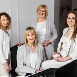 Zespół biura rachunkowego w Siedlcach: cztery kobiety w eleganckich strojach pozują w biurze, uśmiechają się, tworząc profesjonalny i przyjazny wizerunek firmy.