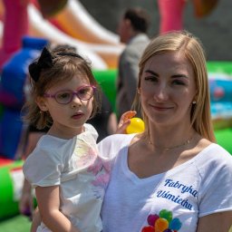 Fabryka Uśmiechu - Uśmiechnięta kobieta w koszulce z napisem 'Fabryka Uśmiechu' trzyma dziewczynkę w okularach na tle dmuchanych atrakcji.