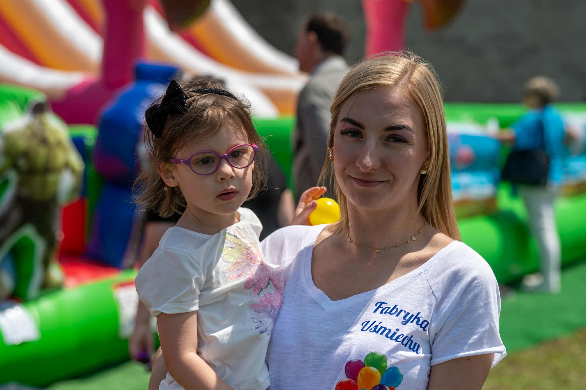 Uśmiechnięta kobieta w koszulce z napisem 'Fabryka Uśmiechu' trzyma dziewczynkę w okularach na tle dmuchanych atrakcji.