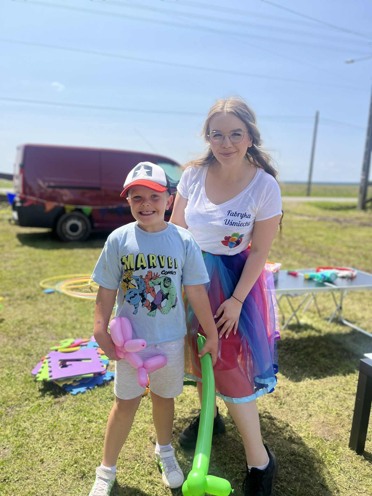Animator w tęczowej spódnicy i chłopiec z balonem-pieskiem na trawie, obok rekwizyty i bus z dekoracją. Logo 'Fabryka Uśmiechu' na koszulce animatorki.