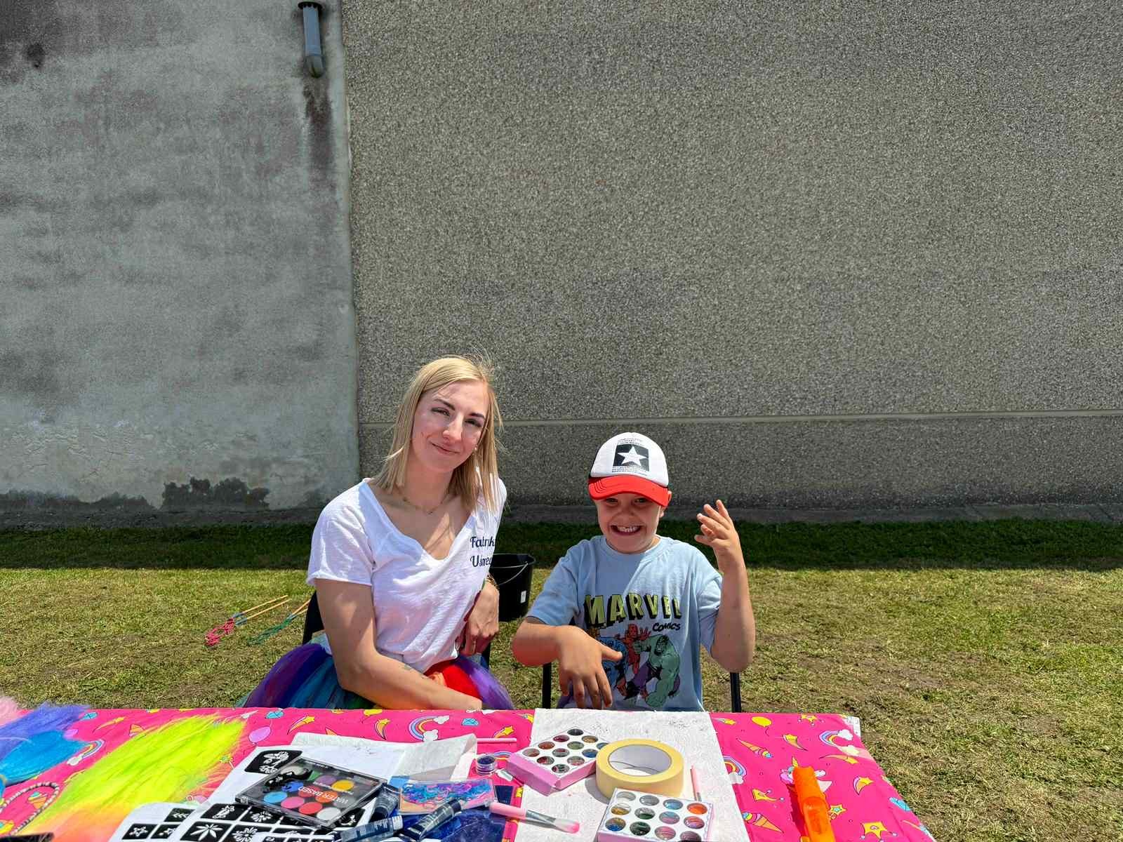 Animator z dzieckiem przy stole z farbami i szablonami do malowania twarzy. Radosna atmosfera, słoneczny dzień, zabawa na świeżym powietrzu.