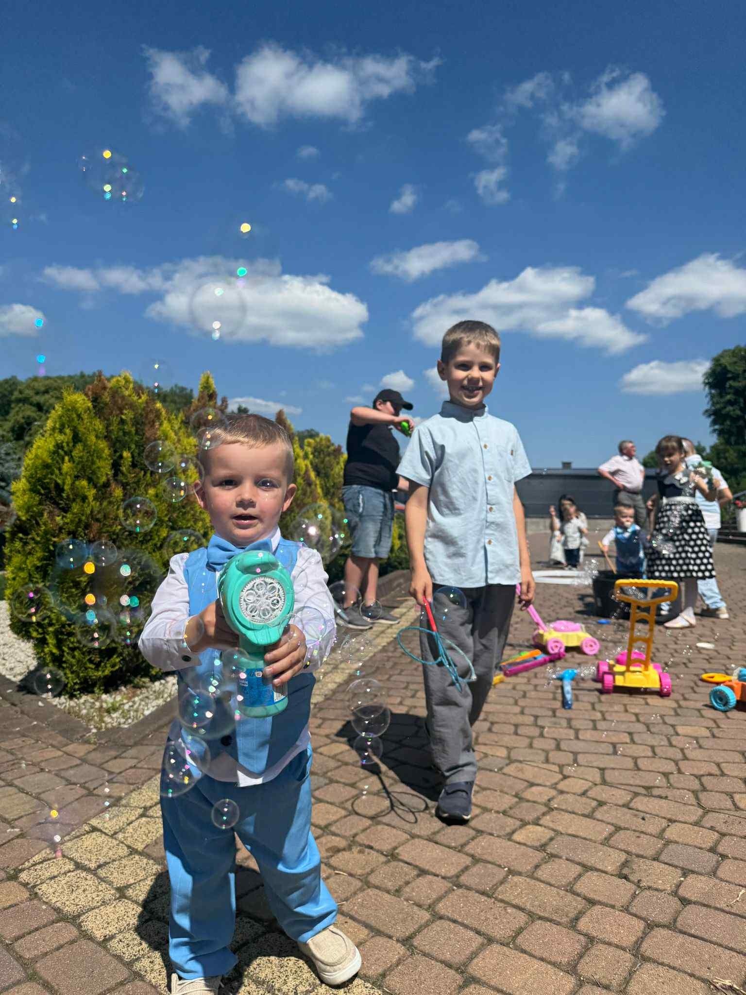 Chłopiec w niebieskim garniturze dmucha bańki mydlane na tle błękitnego nieba i innych dzieci bawiących się na zewnątrz, tworząc radosną atmosferę.